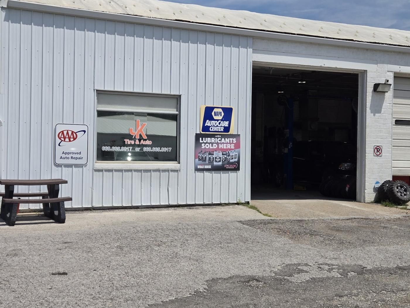 An open service bay beside the shop window with the JK Tire & Auto decal, the AAA Approved Auto Repair sign and the NAPA AutoCare Center sign